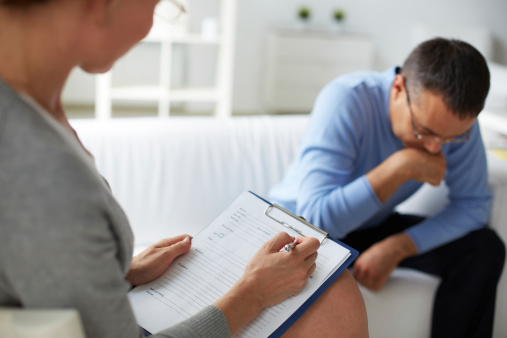 A therapist with a clipboard talks to a man in therapy