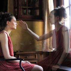 Young adult in red dress with hair pulled back sits somberly in chair with ghostly twin reaching out toward her